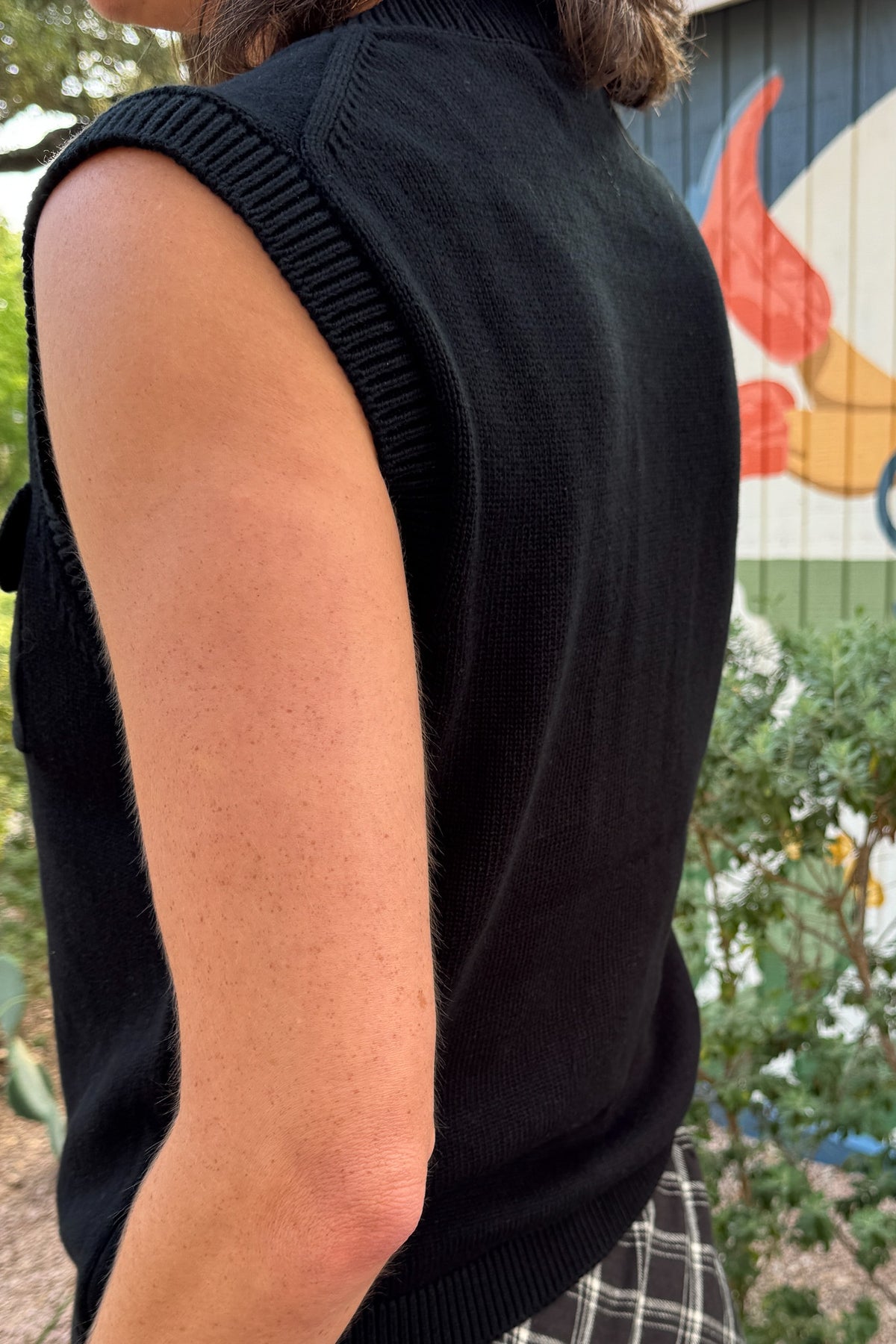 Close-up of black 100% cotton sweater vest with button flap pockets and ribbed armhole detail.