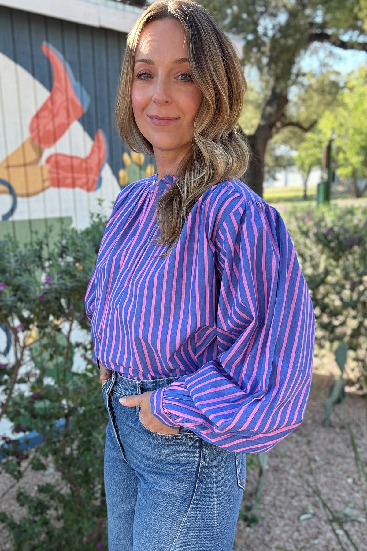 Long sleeve striped blouse for women with bold pink and blue stripes, gathered shoulder detailing, button cuffs, and a feminine tie-neck design.