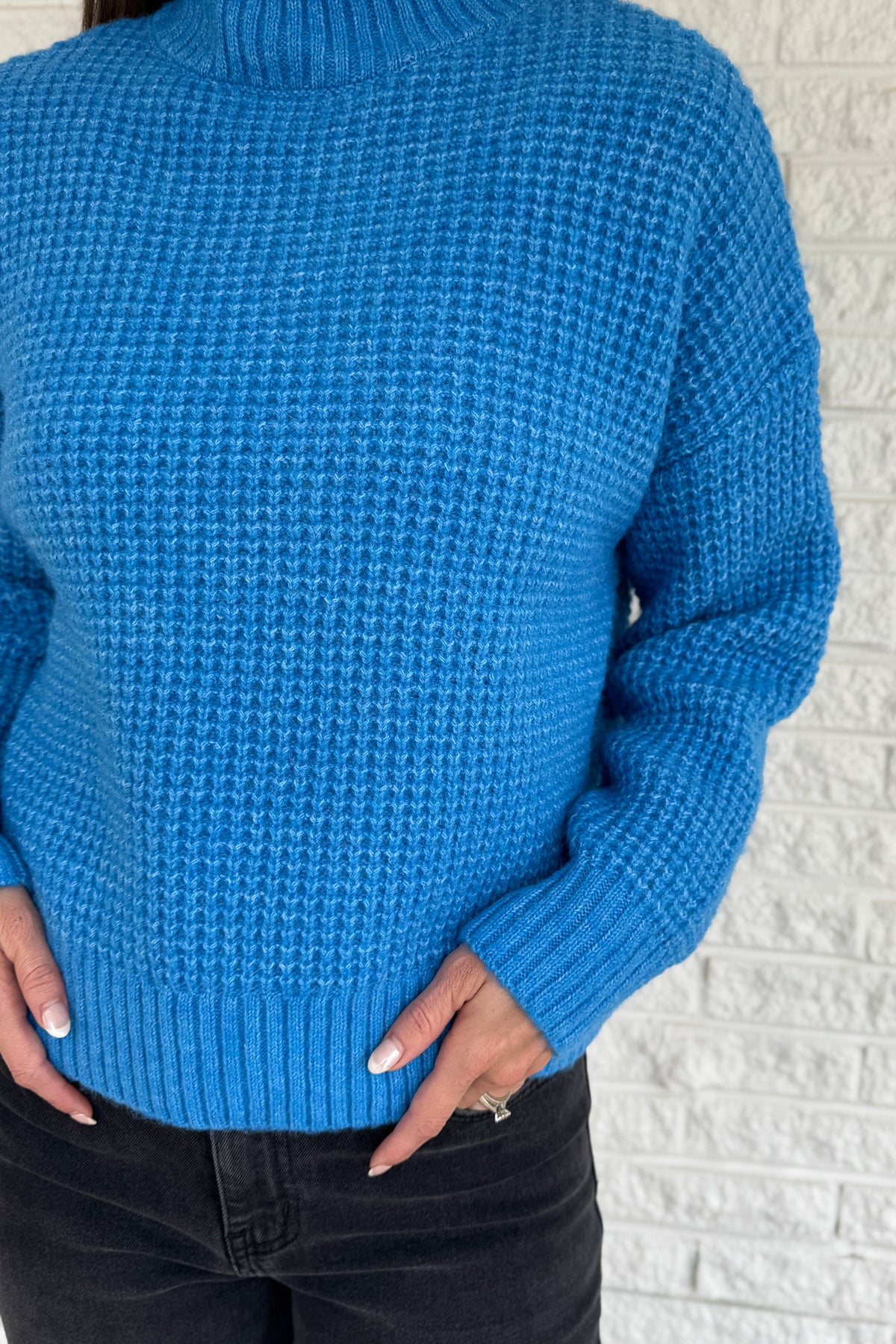 This bold blue oversized sweater brings warmth and style together in one easy piece. Knitted in a soft, textured “square” weave, it features a cozy stand-up collar and ribbed finishes that keep the look clean and structured. Perfect for pairing with denim or a skirt for effortless everyday wear.