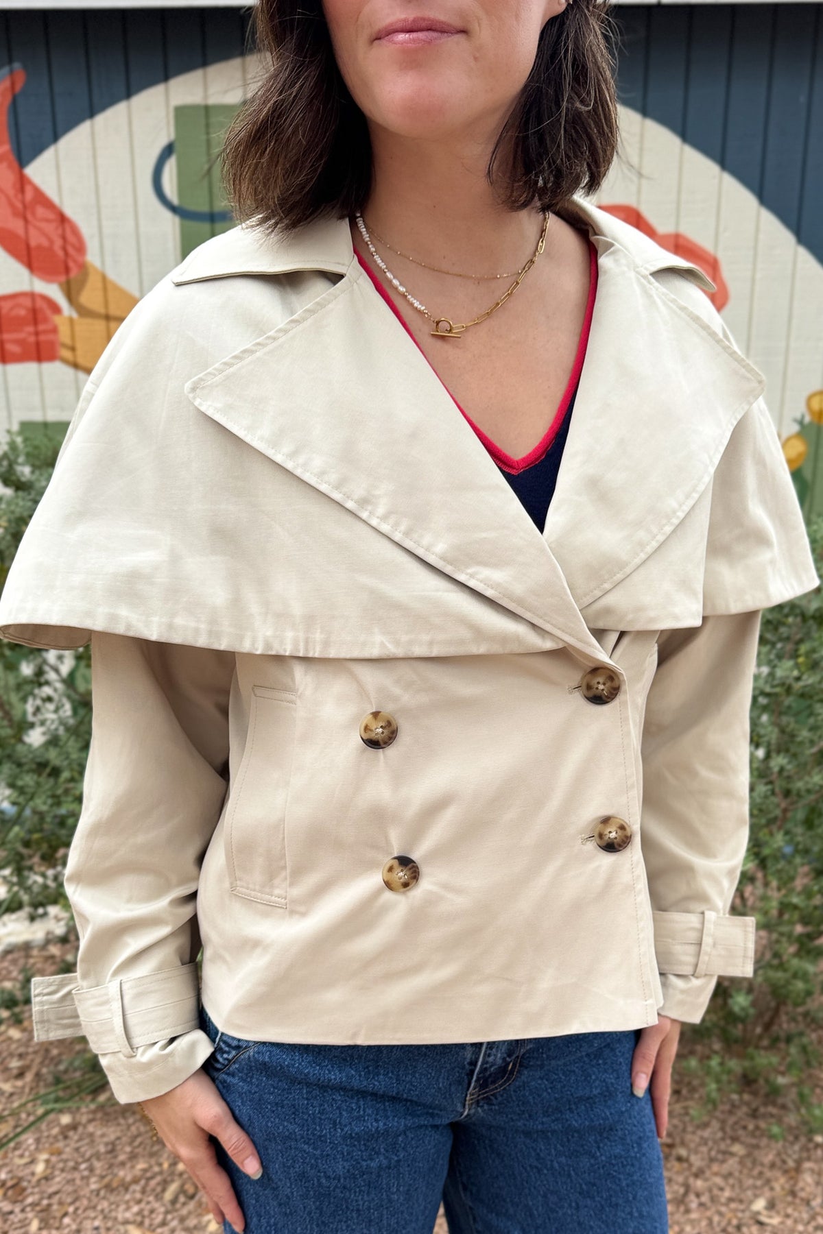Women’s cropped beige trench coat with removable cape overlay, double-breasted buttons, wide lapels and cuff straps at Cotton & Cashmere in Blanco, Texas.