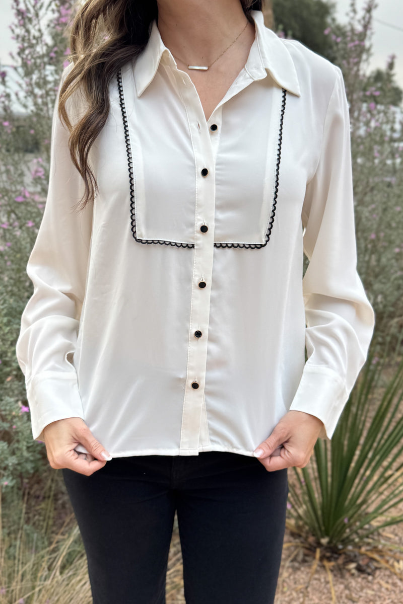A classic button-up with subtle contrast detailing. This blouse features a clean front accented with black scalloped trim and matching buttons for a polished finish. Lightweight with a soft drape, it transitions easily from work to dinner and layers well under blazers or jackets.