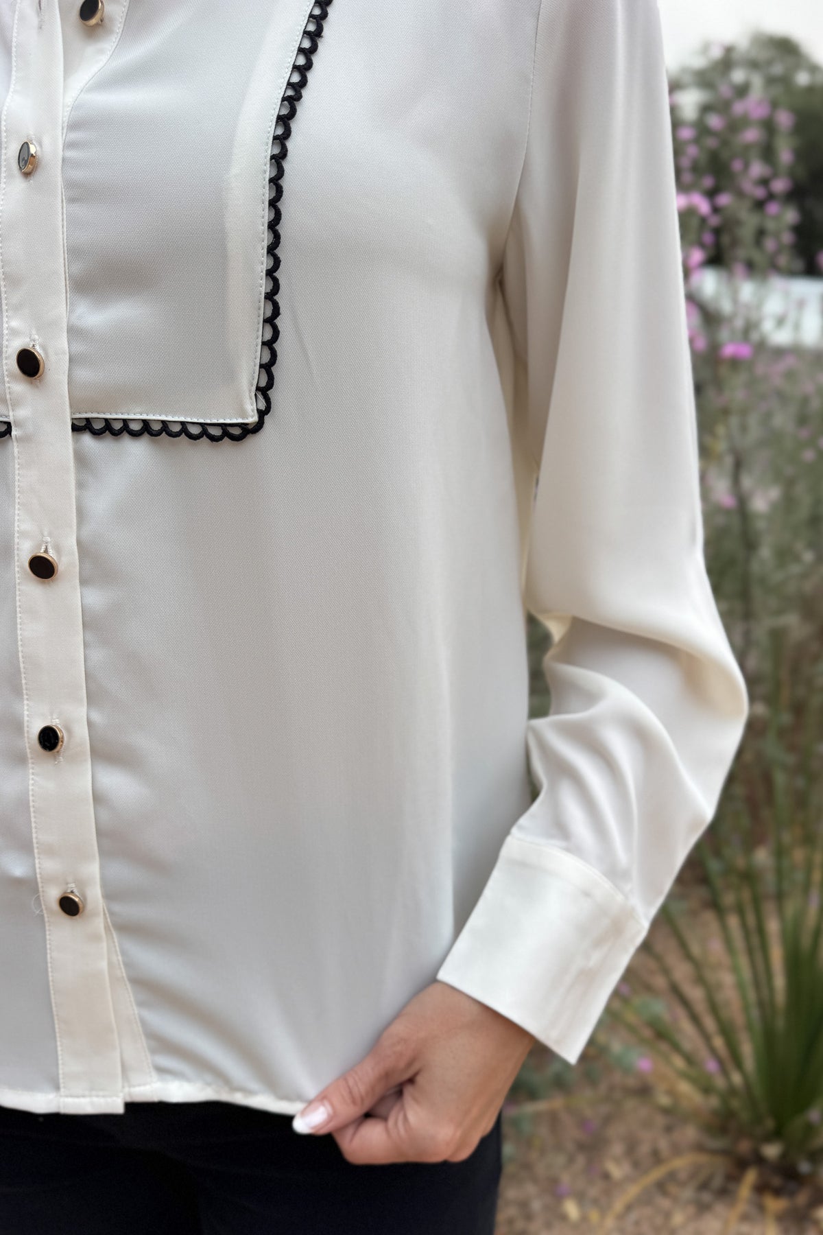 A classic button-up with subtle contrast detailing. This blouse features a clean front accented with black scalloped trim and matching buttons for a polished finish. Lightweight with a soft drape, it transitions easily from work to dinner and layers well under blazers or jackets.
