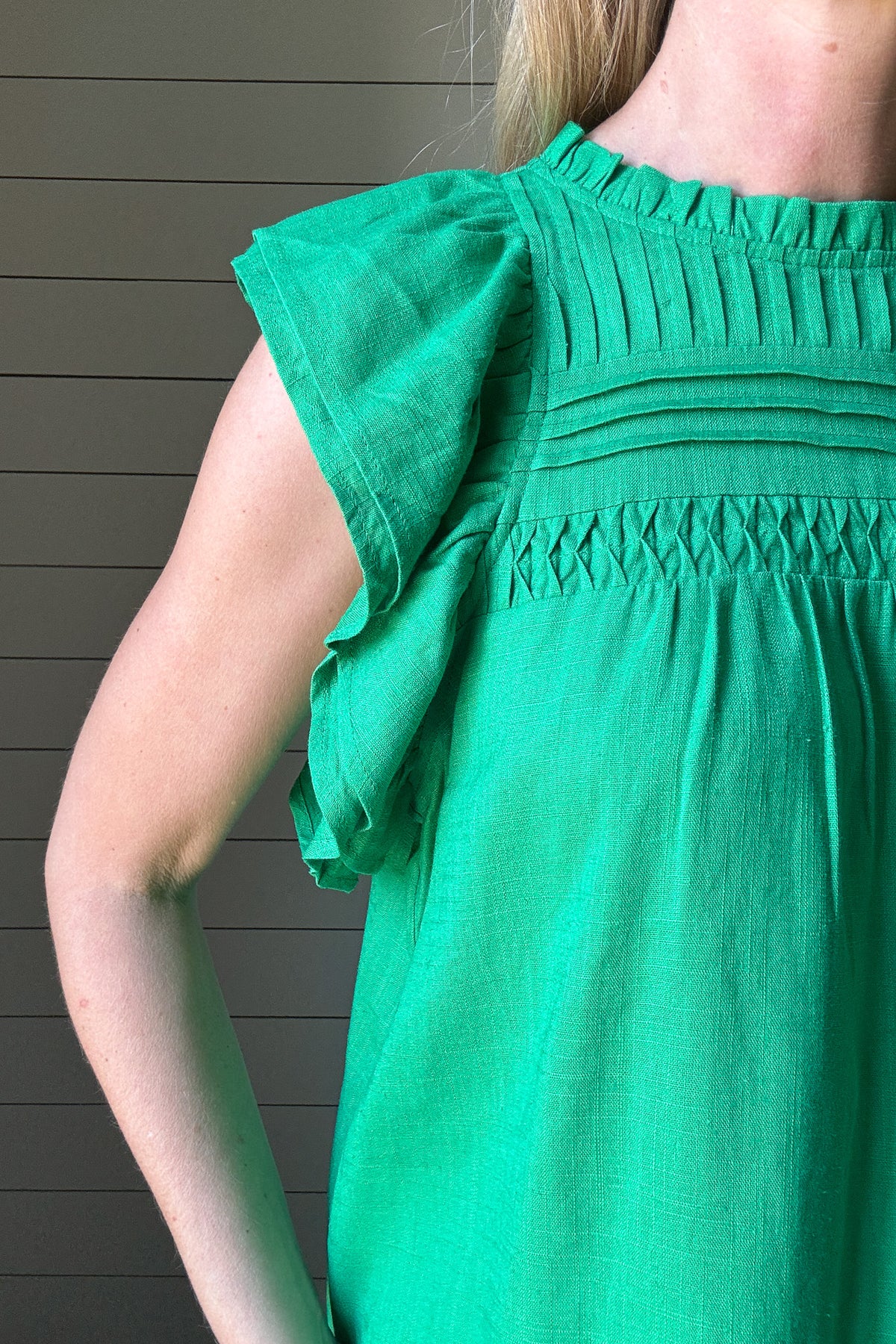 Say hello to your new favorite top. This bright green blouse by THML is all about effortless style with standout details. Made from a woven fabric, it features delicate pleating and smocking across the chest for texture and charm. The flutter sleeves add a soft, feminine touch while keeping it breezy and cool. A high neckline keeps it polished, making it perfect for work or weekend wear. Style it with white denim or tuck it into a skirt, easy either way. It’s a vibrant pop of color for your spring-to-summer