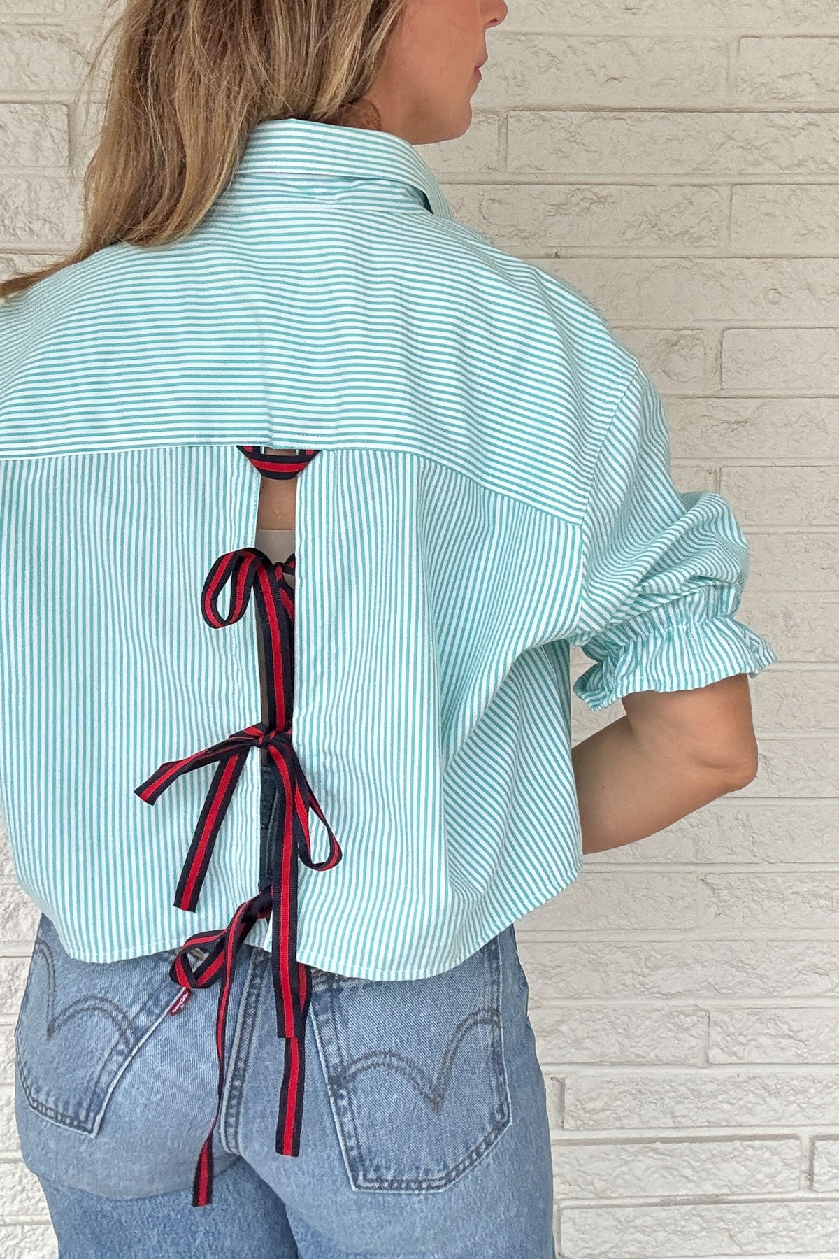 Front photo:
VonVie Harlow aqua and white striped cropped button down shirt with chest pocket, puff sleeves, and ruffled cuffs

Back photo:
Back view of VonVie Harlow aqua striped shirt with open back and red and navy ribbon ties
