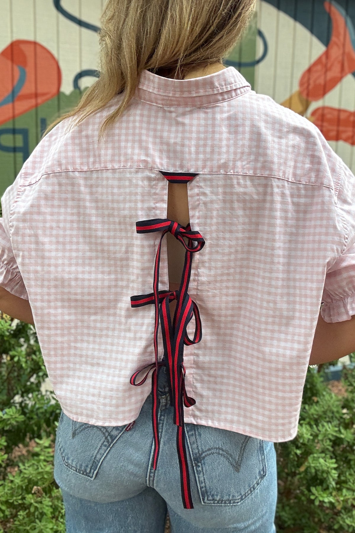 Back photo:
Back view of VonVie Harlow pink gingham cropped shirt with open back and red and navy ribbon ties

Side photo:
Side view of VonVie Harlow pink gingham shirt with puff sleeves, ruffled cuffs, and a relaxed cropped fit