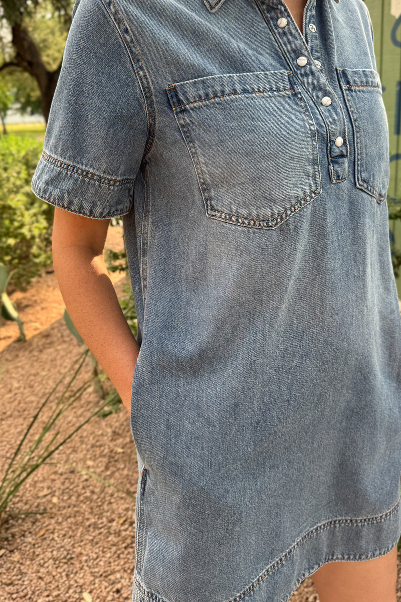 Side view of a women’s light wash denim shirt dress showing the relaxed shape, curved hem and functional side pocket.