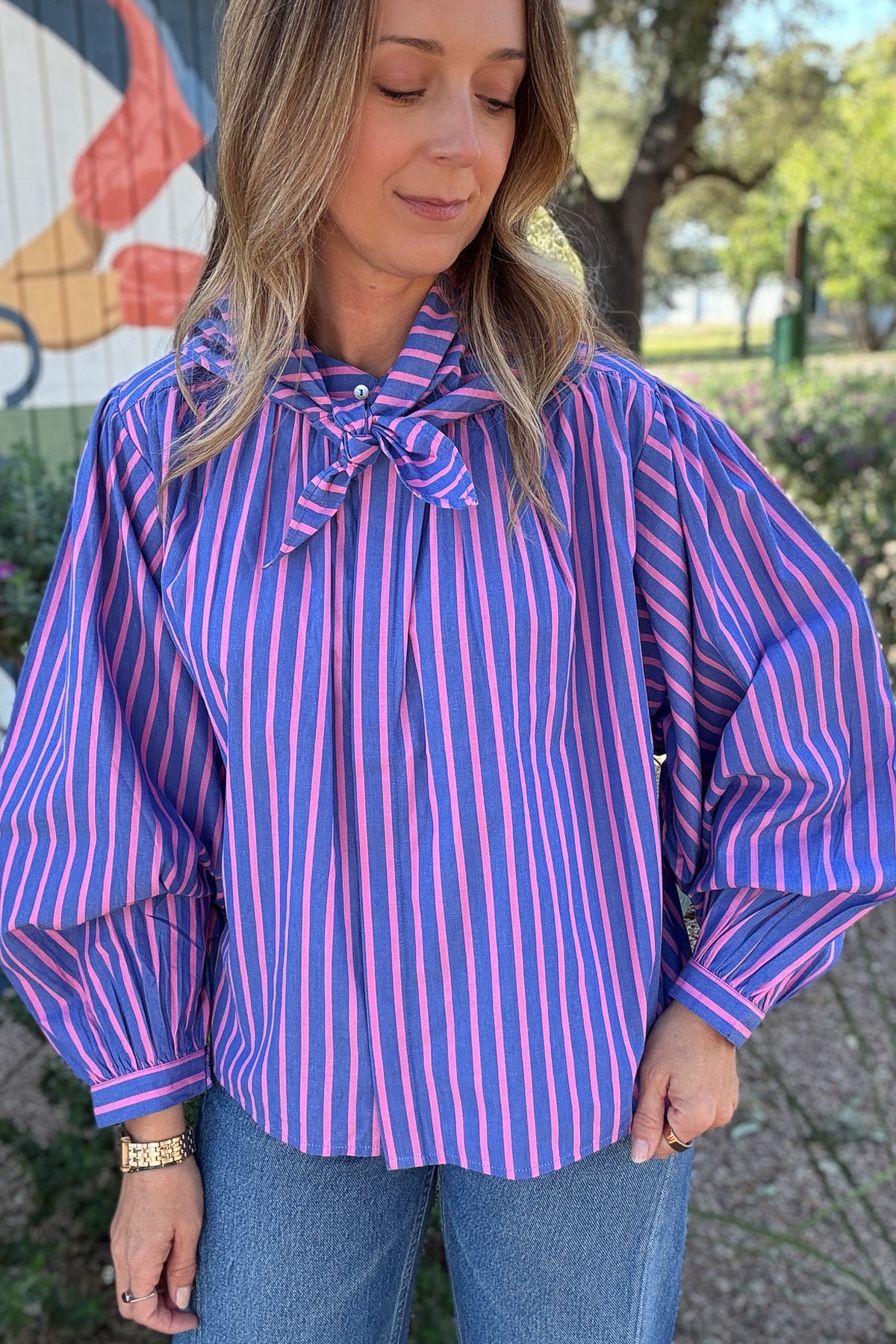 Women's pink and cobalt blue striped blouse with a removable-looking matching scarf tie, gathered shoulders, and billowy sleeves. Styled with classic blue jeans.