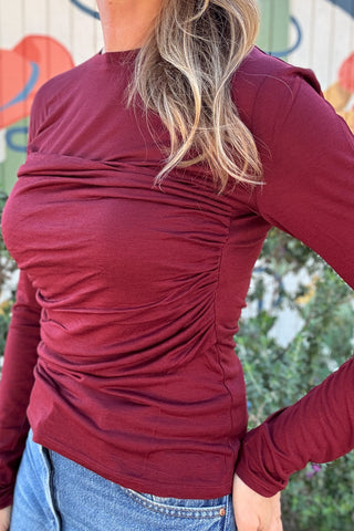 Women's burgundy ruched knit top with a classic round neckline, long sleeves, and flattering gathered detailing. Styled with blue denim at Cotton & Cashmere in Blanco, Texas.