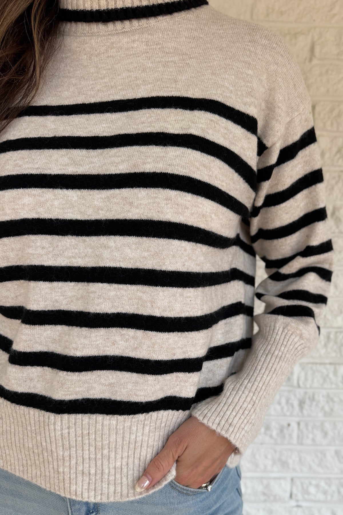 Close-up of a person wearing a beige sweater with black horizontal stripes against a light brick wall.
