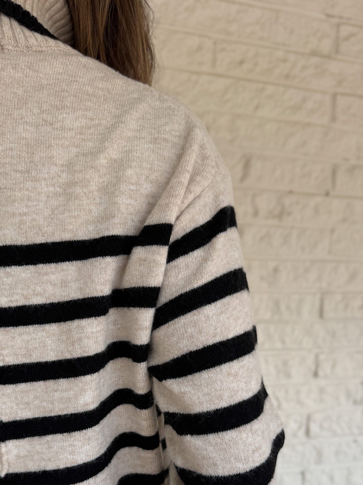 Close-up of a person wearing a striped sweater with black and beige pattern against a light brick wall.