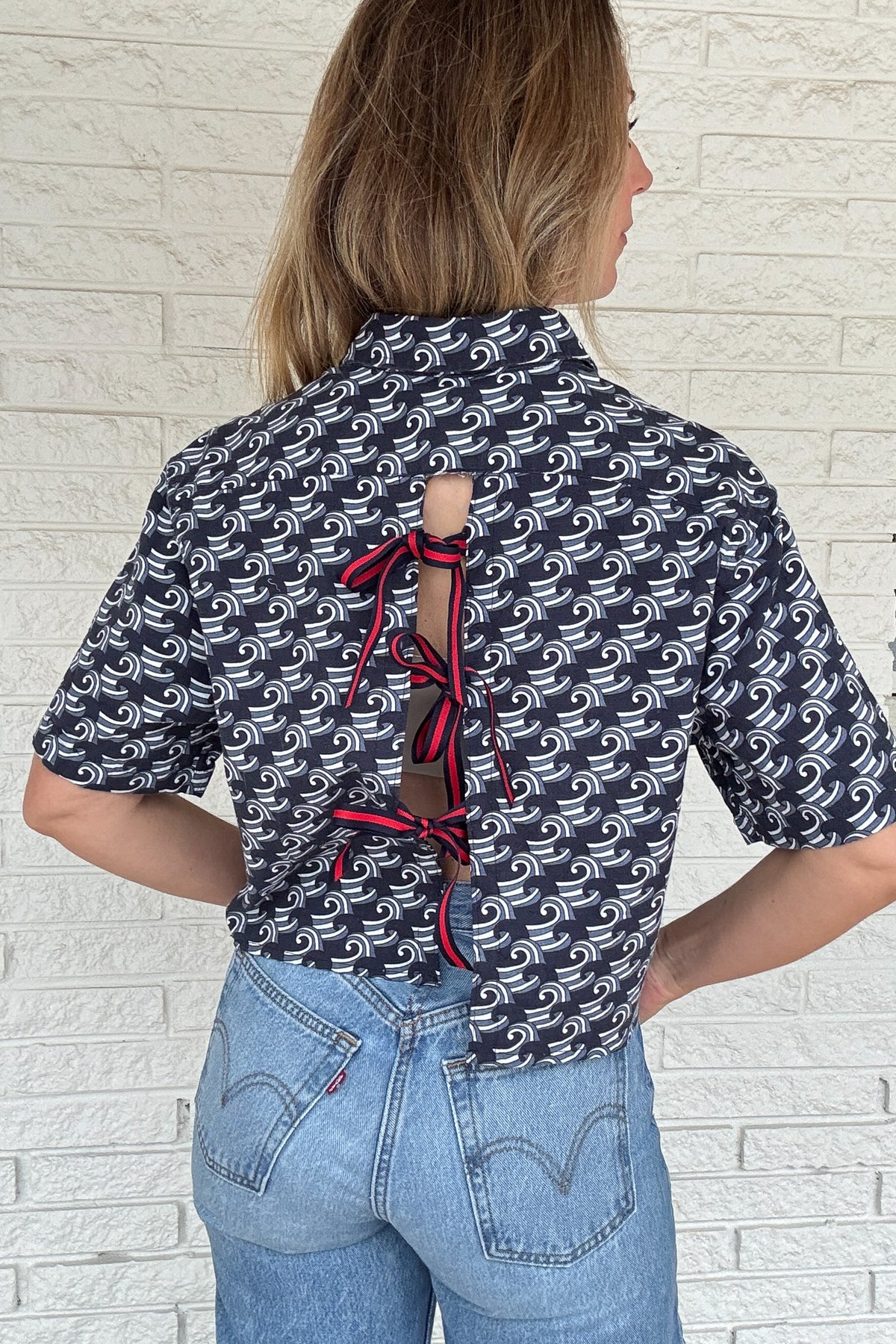 Front photo:
VonVie Harlow navy and white wave print cropped button down shirt with relaxed elbow length sleeves

Back photo:
Back view of VonVie Harlow navy wave print shirt with open back and red and navy ribbon ties