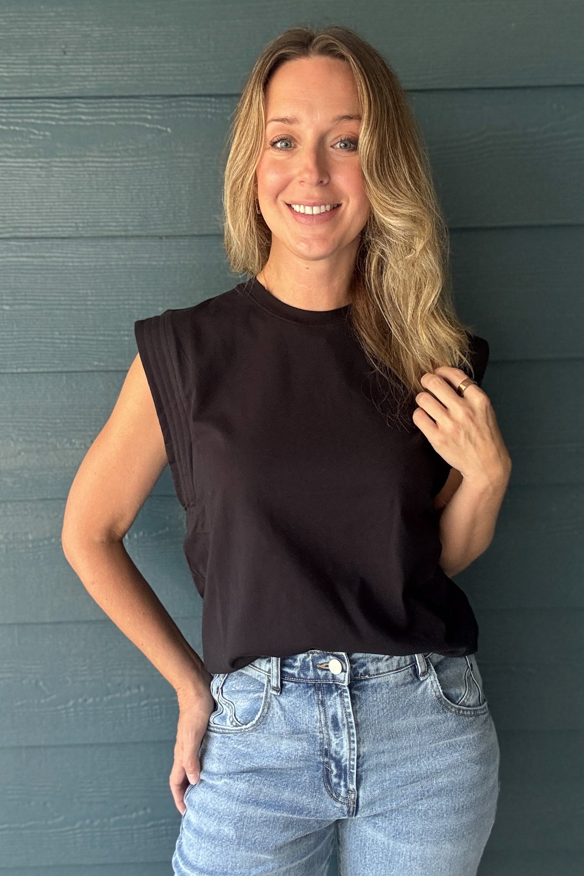 Women’s black structured sleeveless top with padded shoulders, crew neckline and relaxed fit, styled with light wash jeans.