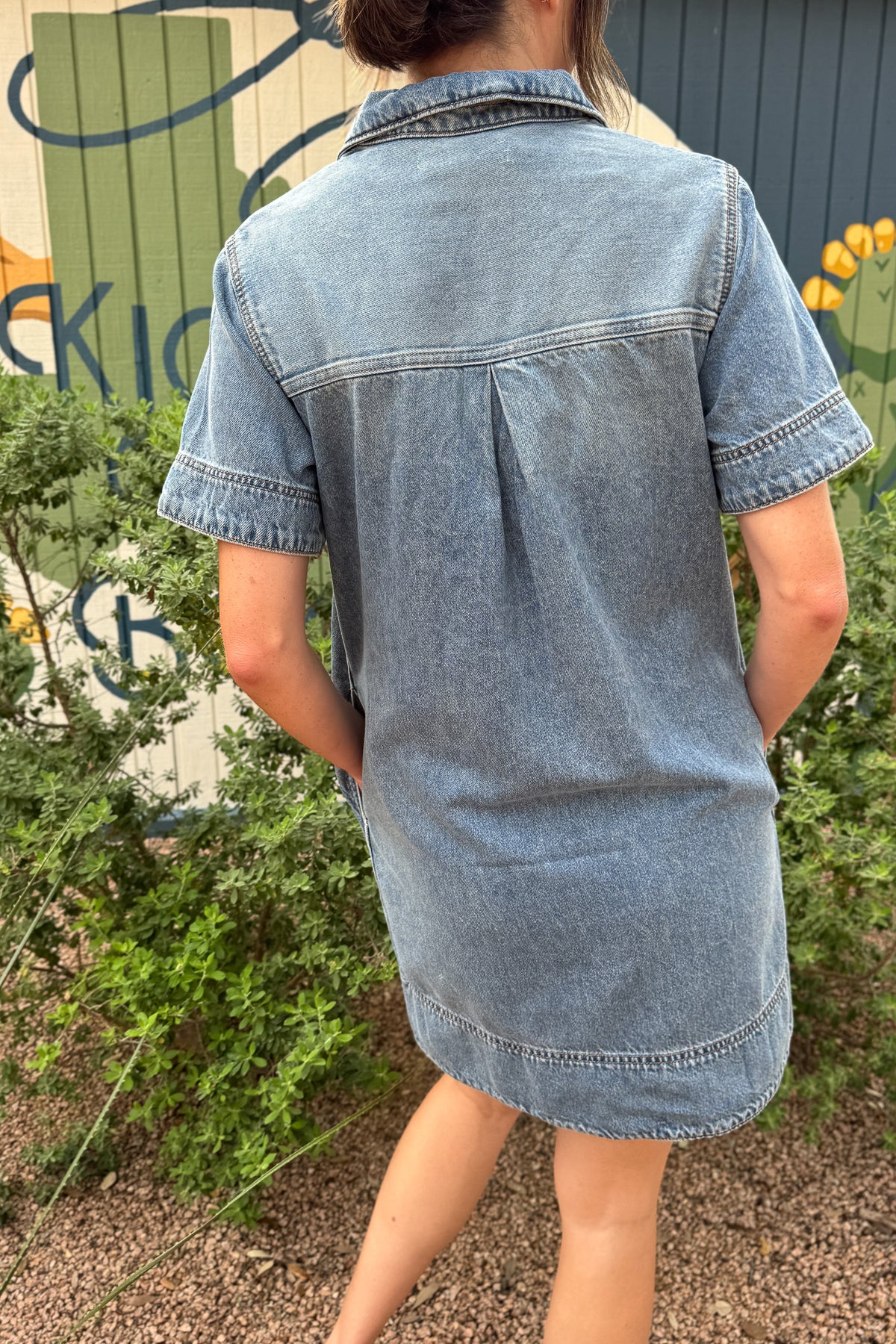 Back view of a women’s light wash denim shirt dress with short sleeves, a back yoke and center pleat for a relaxed fit.