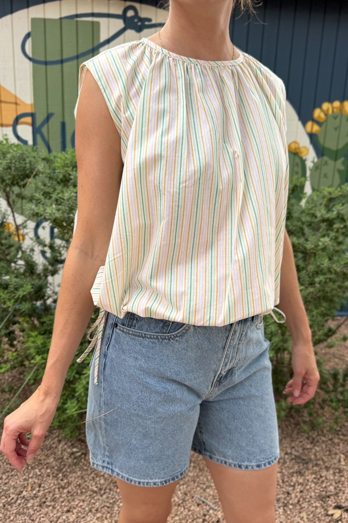 Back view of a women’s pastel striped sleeveless top with a relaxed fit, gathered neckline, button closure and adjustable side ties.