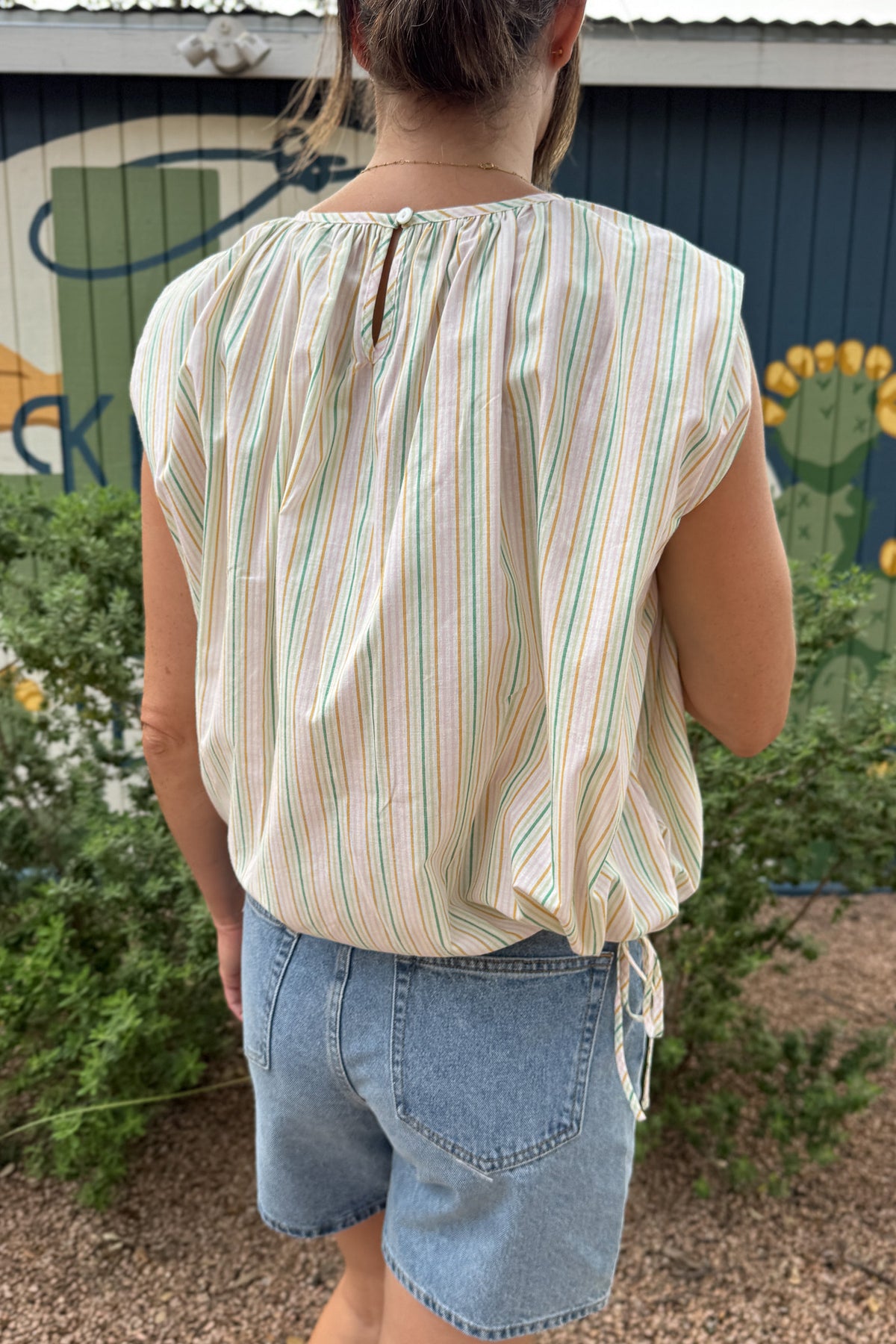 Women’s pastel striped sleeveless top with a gathered round neckline, relaxed fit and adjustable side ties, styled with denim shorts.